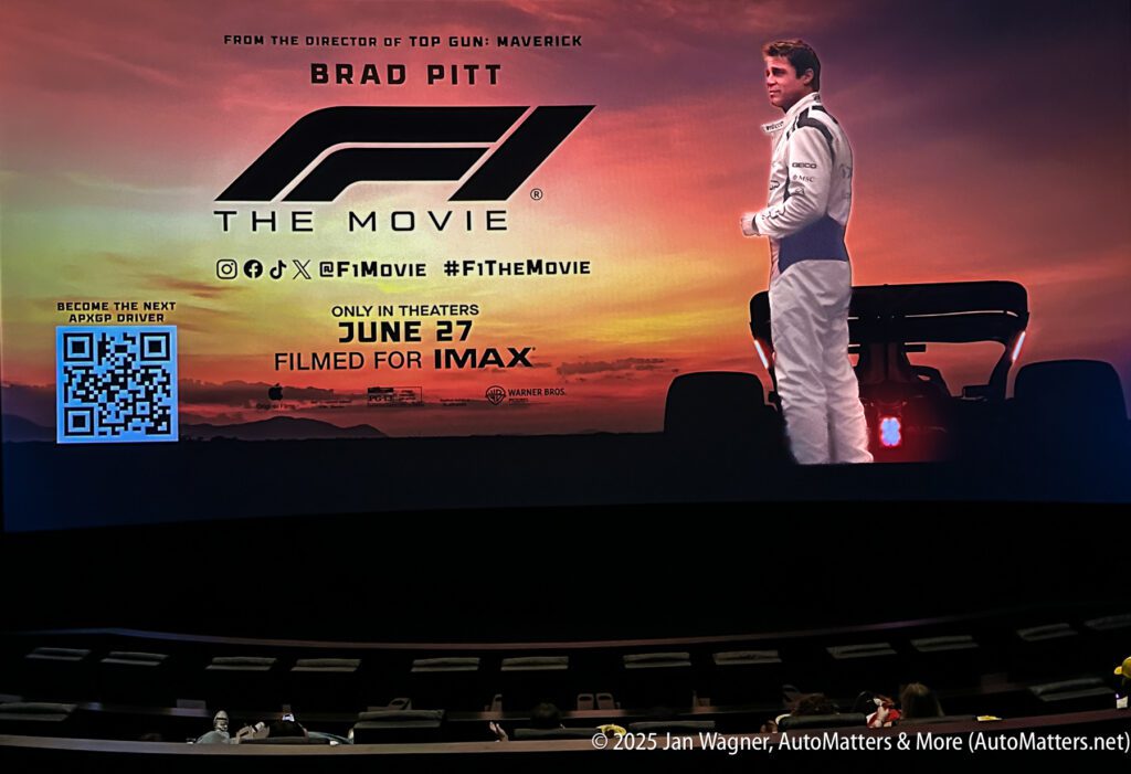 Large movie theater screen displays promo for "F1: The Movie" featuring a man in a racing suit, film details, QR code, and seating silhouettes in the foreground.