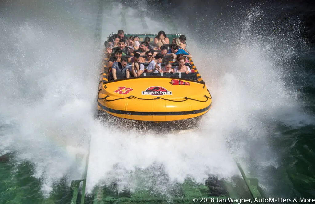 A yellow Jurassic Park ride boat splashes into water, soaking passengers with spray as they brace themselves.
