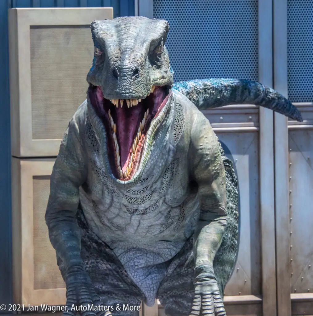A realistic animatronic dinosaur with its mouth open stands in front of metallic doors.