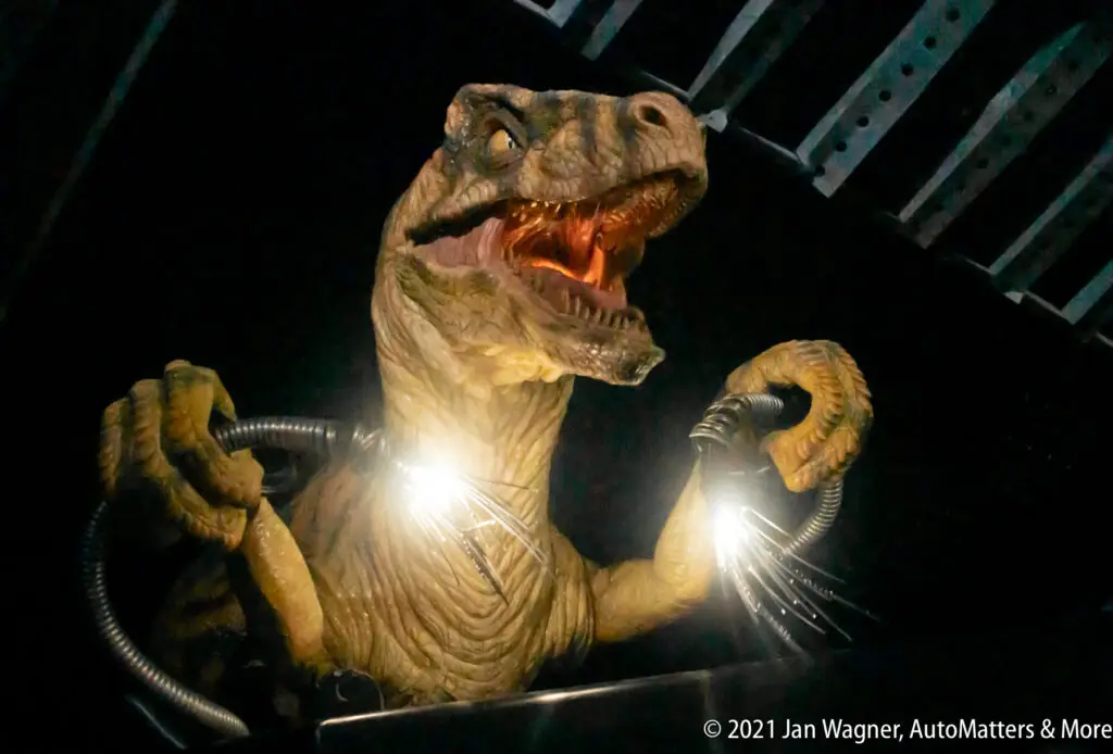 A model of a dinosaur with an open mouth holds cables emitting sparks in a dark setting.