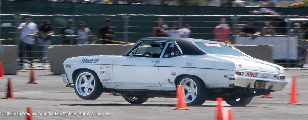 A white classic muscle car with racing stickers drifts around orange cones at an outdoor event, with spectators watching behind a barrier.