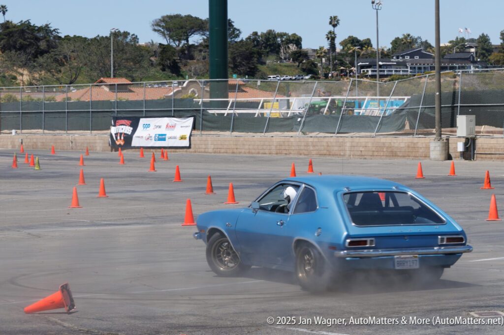 A blue classic car navigates an autocross course marked by orange cones in a parking lot, with tire smoke visible behind the vehicle.