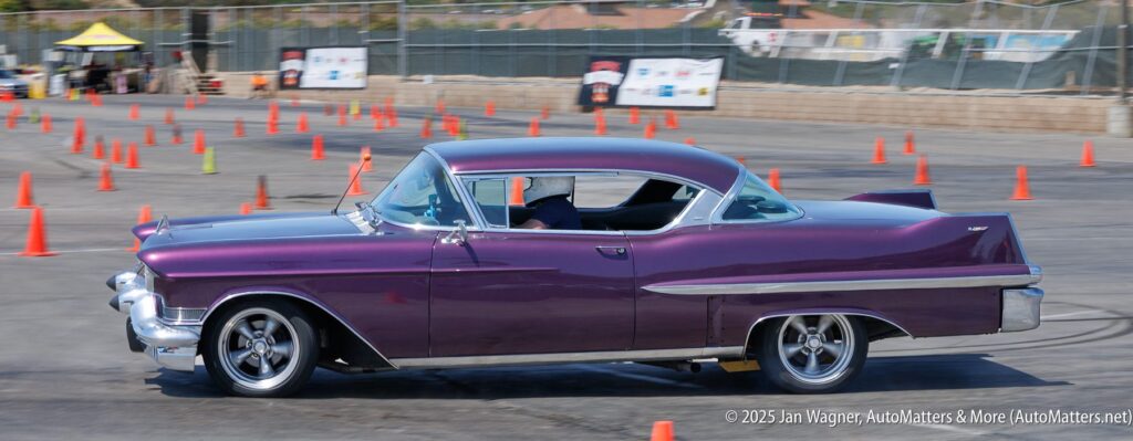 A purple vintage car drives through an autocross course marked by orange traffic cones in a parking lot.