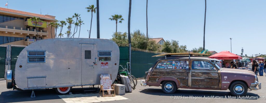 A vintage station wagon with surfboards on top is parked beside a retro teardrop camper trailer at an outdoor event under palm trees.