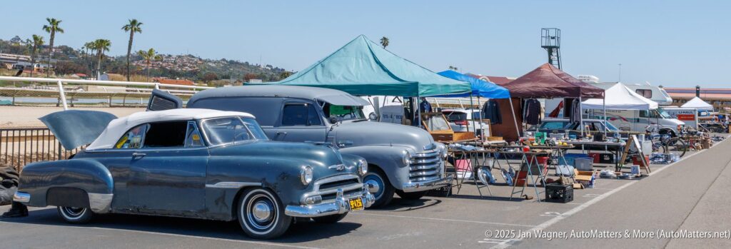 Classic cars parked beside vendor tents displaying various items at an outdoor swap meet under clear skies.