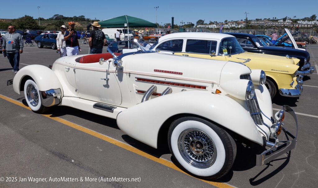 A white vintage Auburn Speedster convertible is parked at a car show, with other classic cars and people visible in the background.