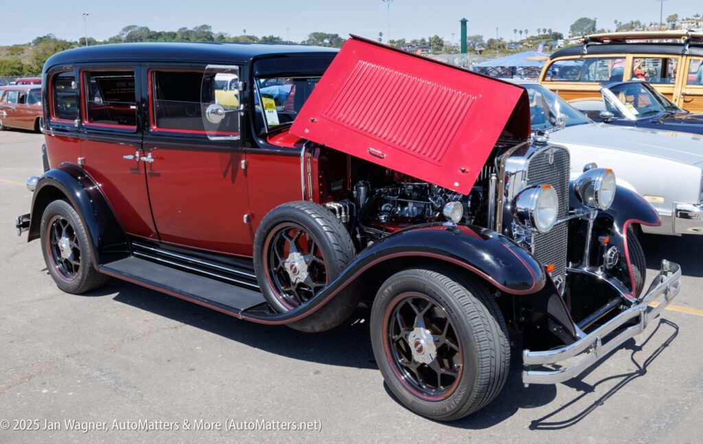 A vintage maroon and black classic car with its hood open is parked at an outdoor car show.