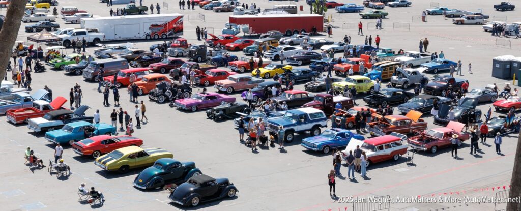 A large outdoor car show features numerous vintage cars of various colors, with attendees walking and gathering around vehicles in a spacious parking lot.