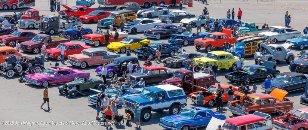 A large outdoor car show features numerous classic cars, hot rods, and trucks parked in rows, with attendees walking around and taking photos.