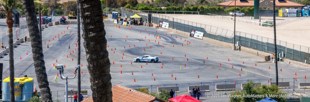 A white sports car maneuvers through an autocross course marked by orange traffic cones in a large, outdoor paved lot.