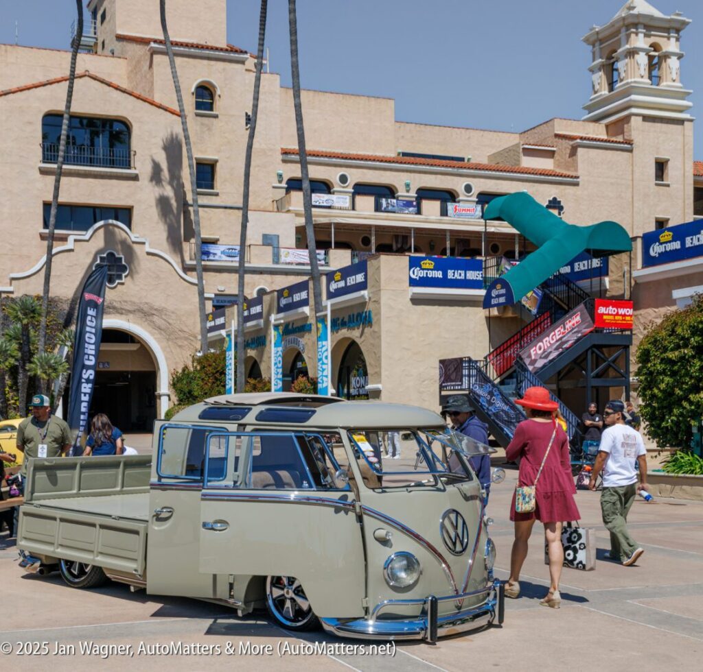 A vintage Volkswagen van is parked at an outdoor event in front of a large building, with people walking and various banners and displays visible.