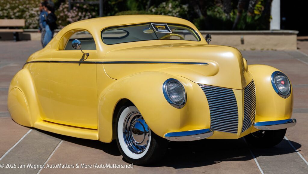 A vintage custom yellow coupe with chrome details and whitewall tires is parked outdoors on a sunny day.