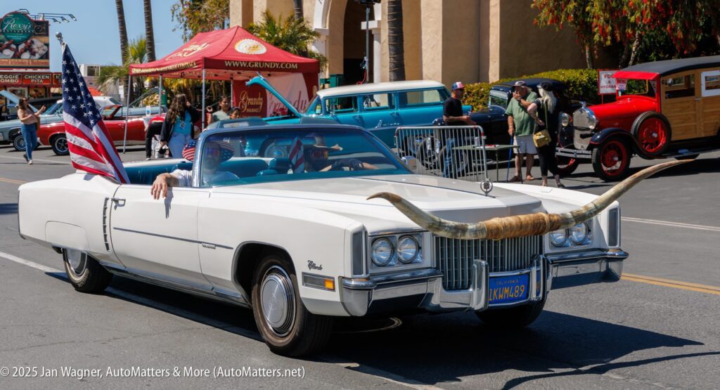 A white vintage convertible with large horns mounted on the front grille drives in a parade, decorated with two American flags.