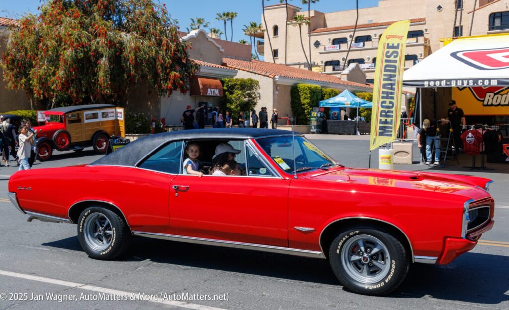 A bright red classic Pontiac GTO with a black vinyl roof drives past vendor booths at an outdoor car show, with two children in the back seat and an adult driver.