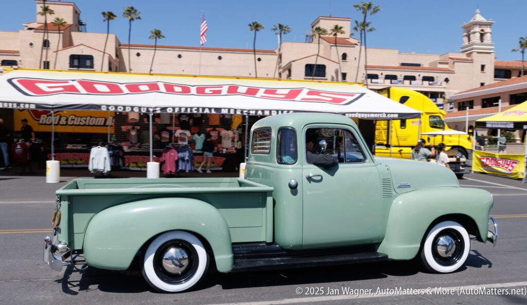 A vintage light green pickup truck drives past vendor tents and a Goodguys banner at an outdoor car show.