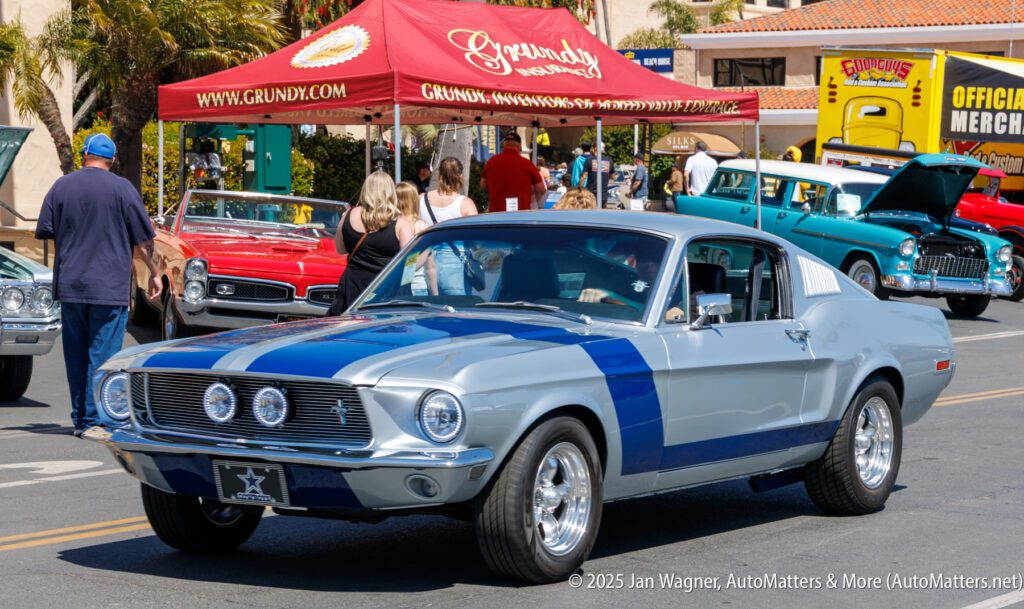 A classic silver and blue Ford Mustang is parked at a car show, with people and other vintage cars visible in the background.