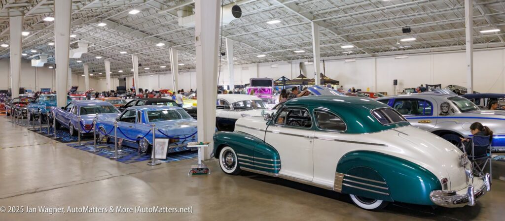 Row of classic cars on display inside a large, well-lit exhibition hall with people viewing the vehicles.
