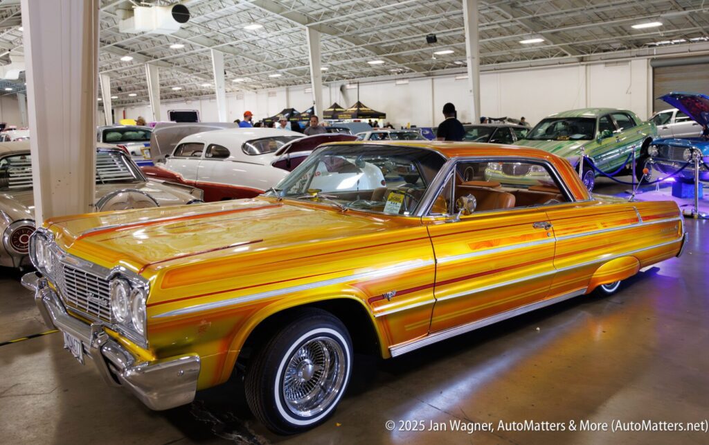 A customized yellow and orange lowrider car with chrome detailing is on display indoors at a car show, surrounded by other classic vehicles.