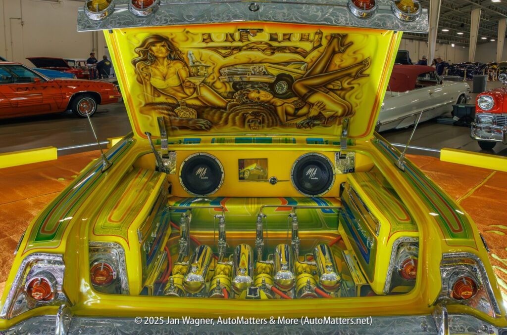 Customized classic car trunk features vibrant yellow and green paint, detailed mural art on the lid, chrome hydraulic pumps, and large speakers in an indoor car show setting.