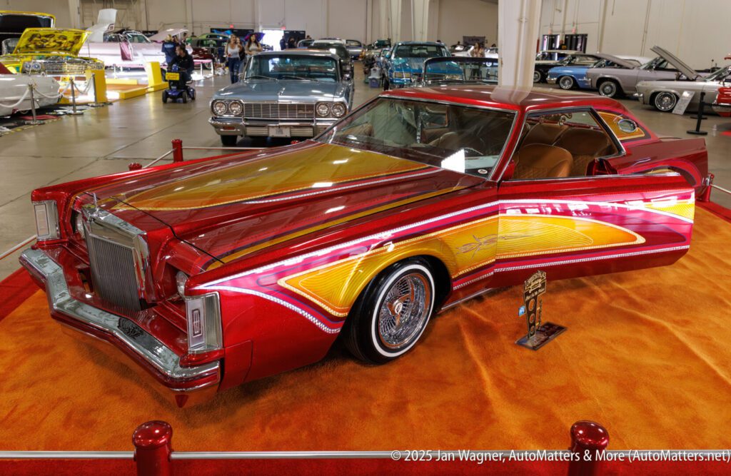 Customized classic car with intricate red, yellow, and gold paintwork displayed indoors on an orange carpet, surrounded by other vintage cars.