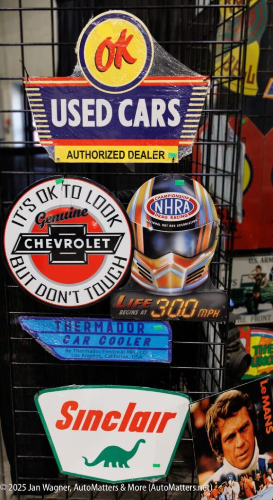 A display of vintage signs includes “OK Used Cars,” Chevrolet, NHRA, Sinclair, and a ThermaDor car cooler, mounted on a metal grid.