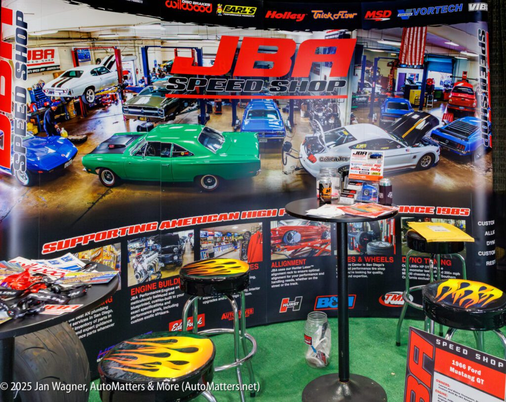 JBA Speed Shop exhibit booth with car posters, display tables, flame-patterned stools, and automotive product brochures and parts on display.