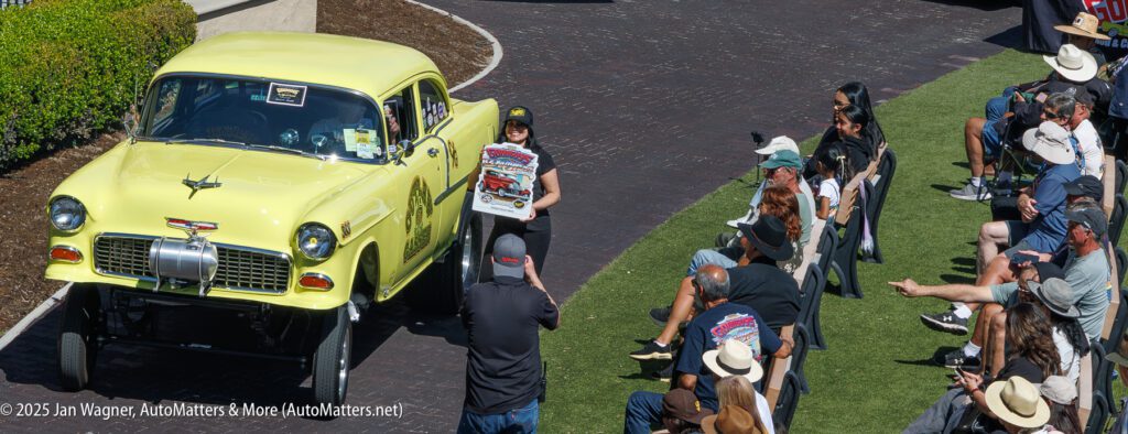 A woman stands in front of a yellow classic car holding a poster, as photographers and spectators watch at an outdoor event.