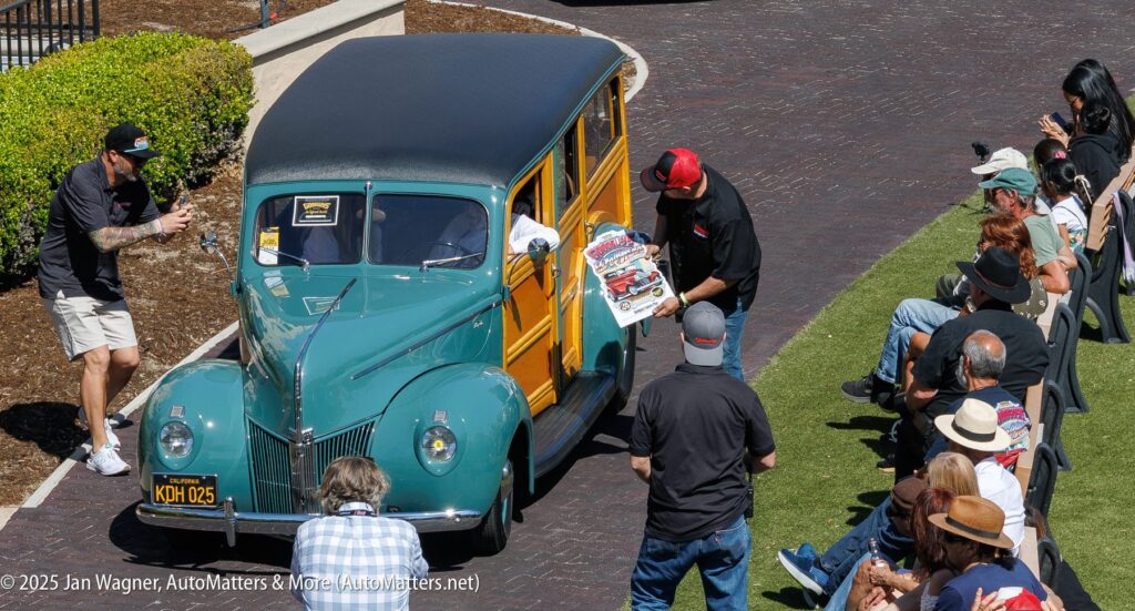 A man in a black shirt presents a framed poster next to a green vintage wood-paneled car in front of an outdoor crowd at an auto show.