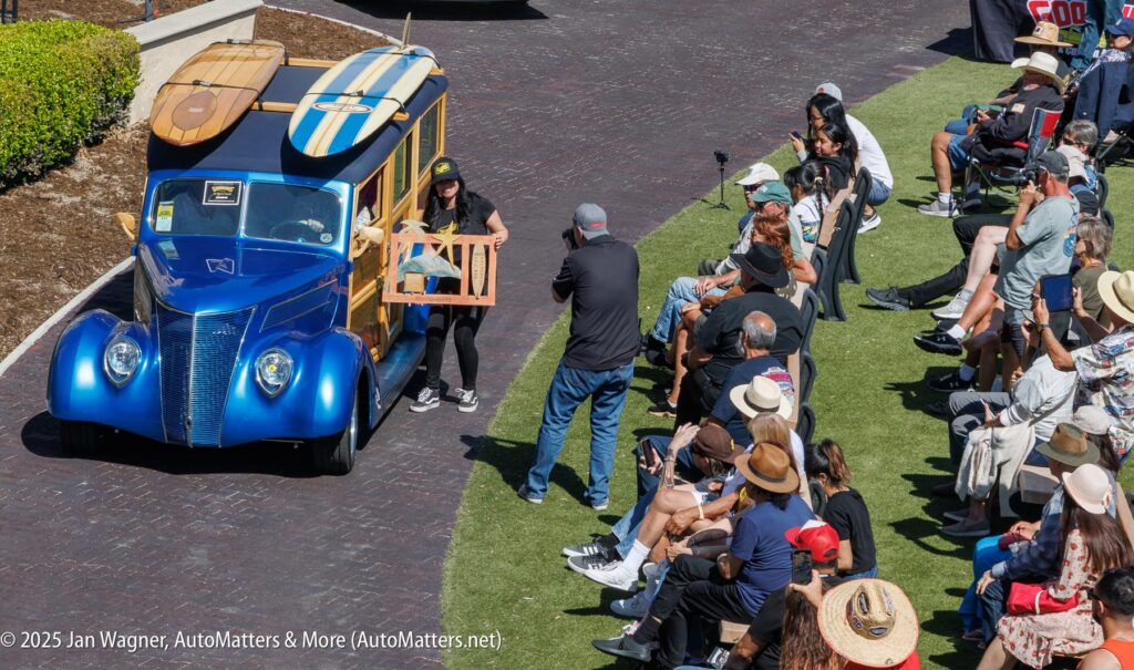 A blue vintage car with surfboards on top is displayed before an audience seated on both sides at an outdoor event.