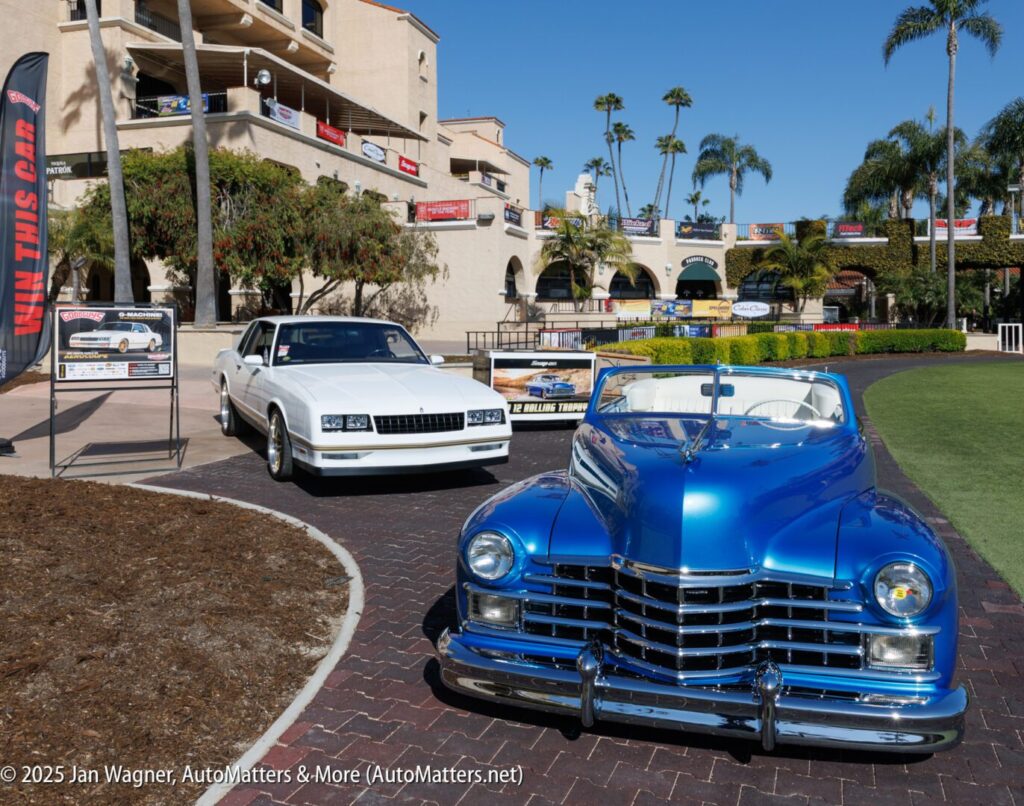 A shiny blue classic convertible and a white coupe are parked outdoors on a brick path near a multi-story building with palm trees and banners in the background.