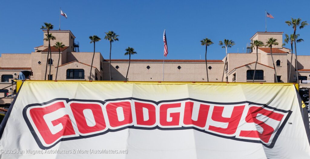 A large "Goodguys" banner is displayed in front of a tan building with palm trees and American flags in the background.
