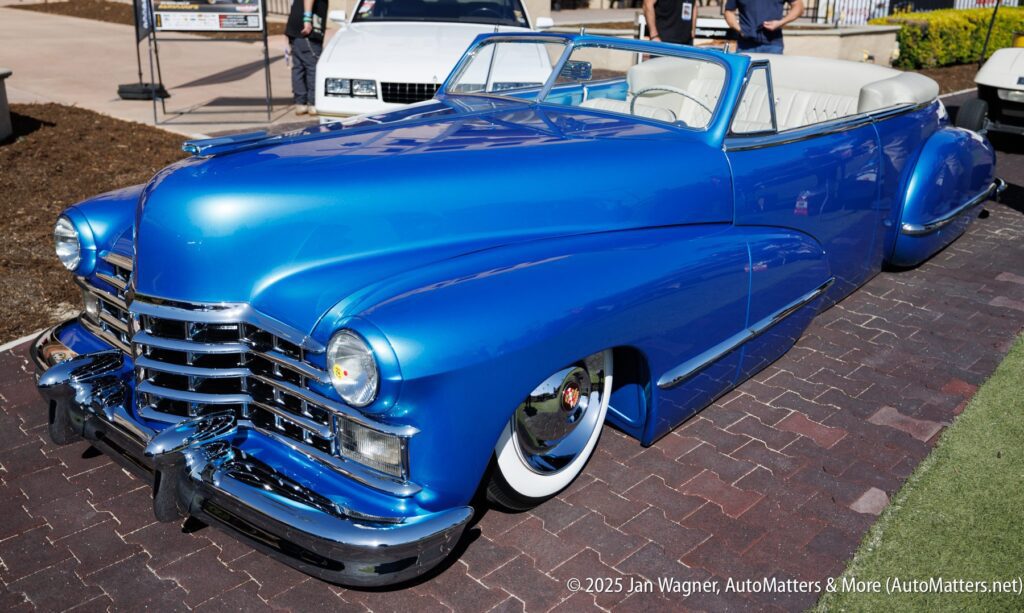 A bright blue classic convertible car with a white interior is parked on a brick surface at an outdoor car show.