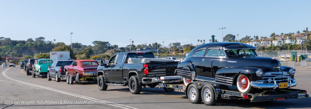 A line of trucks towing classic cars on trailers is parked along a paved road with trees and buildings in the background.