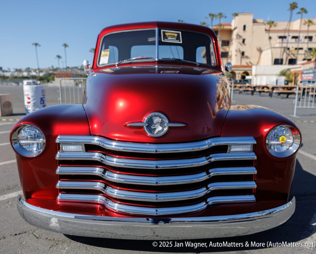 Front view of a shiny red vintage Chevrolet truck parked outdoors at a car show, with palm trees and buildings visible in the background.