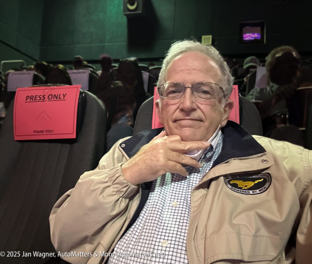 An older man with glasses sits in a movie theater seat marked "Press Only," wearing a beige jacket and checked shirt. Other people are seated in the background.