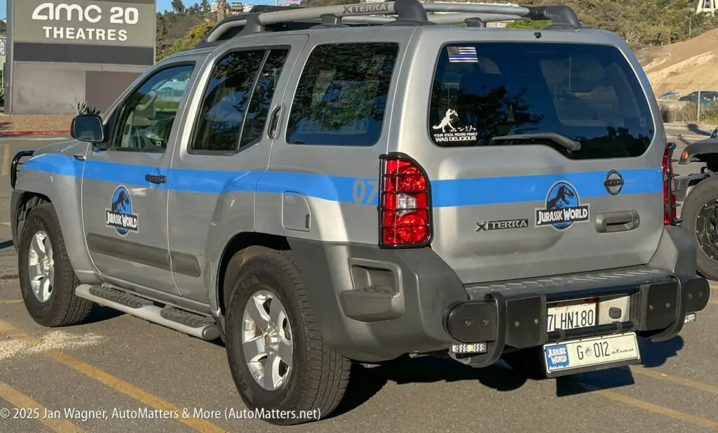 A gray Nissan Xterra SUV with "Jurassic World" decals is parked in a lot near an AMC movie theater. The vehicle displays California license plates.