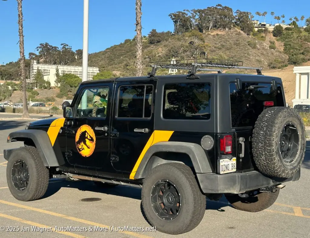 A black Jeep with yellow accents and a Jurassic Park logo is parked outdoors on a sunny day, with trees and buildings in the background.