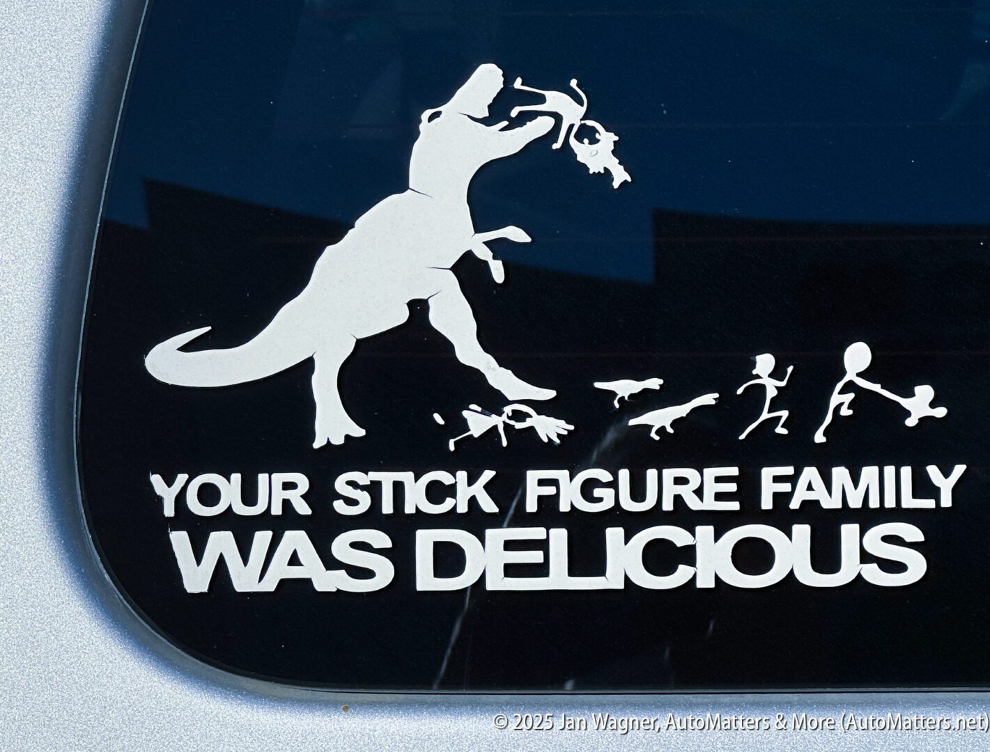 White decal on a car window shows a dinosaur eating stick figure people, with the text "YOUR STICK FIGURE FAMILY WAS DELICIOUS.
