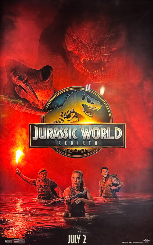 Movie poster for "Jurassic World: Rebirth" showing three people in water with a flare and weapons, a dinosaur logo in the center, and a large dinosaur looming in the background. Release date: July 2.