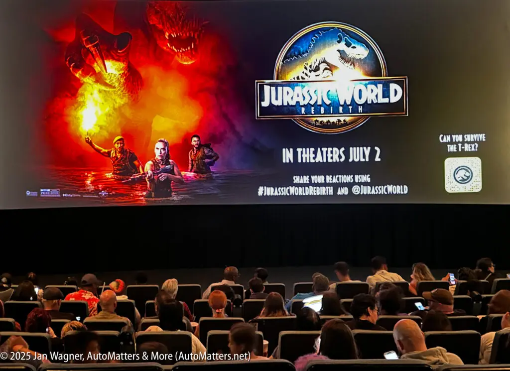 Audience seated in a theater facing a large screen displaying a "Jurassic World Rebirth" movie poster with release date, social media hashtags, and images of dinosaurs and characters.