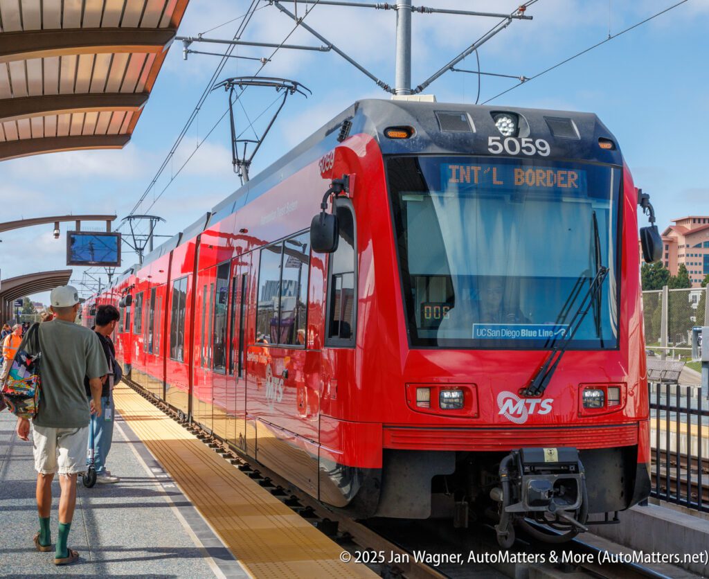 A red San Diego Trolley train labeled "IN'TL BORDER" is stopped at a station platform as passengers walk nearby.