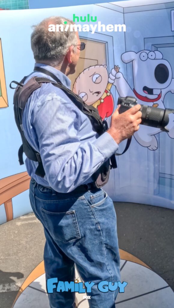 A man in blue jeans and a vest holds a camera in front of a "Family Guy" photo backdrop promoting Hulu's Ani-Mayhem event.