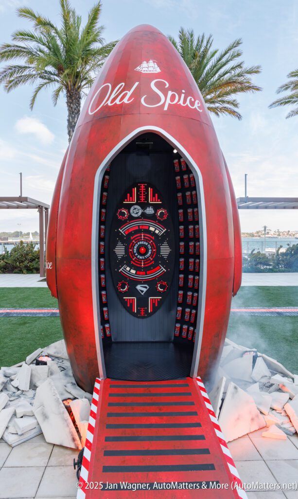 A large red Old Spice-themed rocket-shaped structure stands upright with an open door, revealing a control panel inside; surrounded by palm trees and concrete debris.