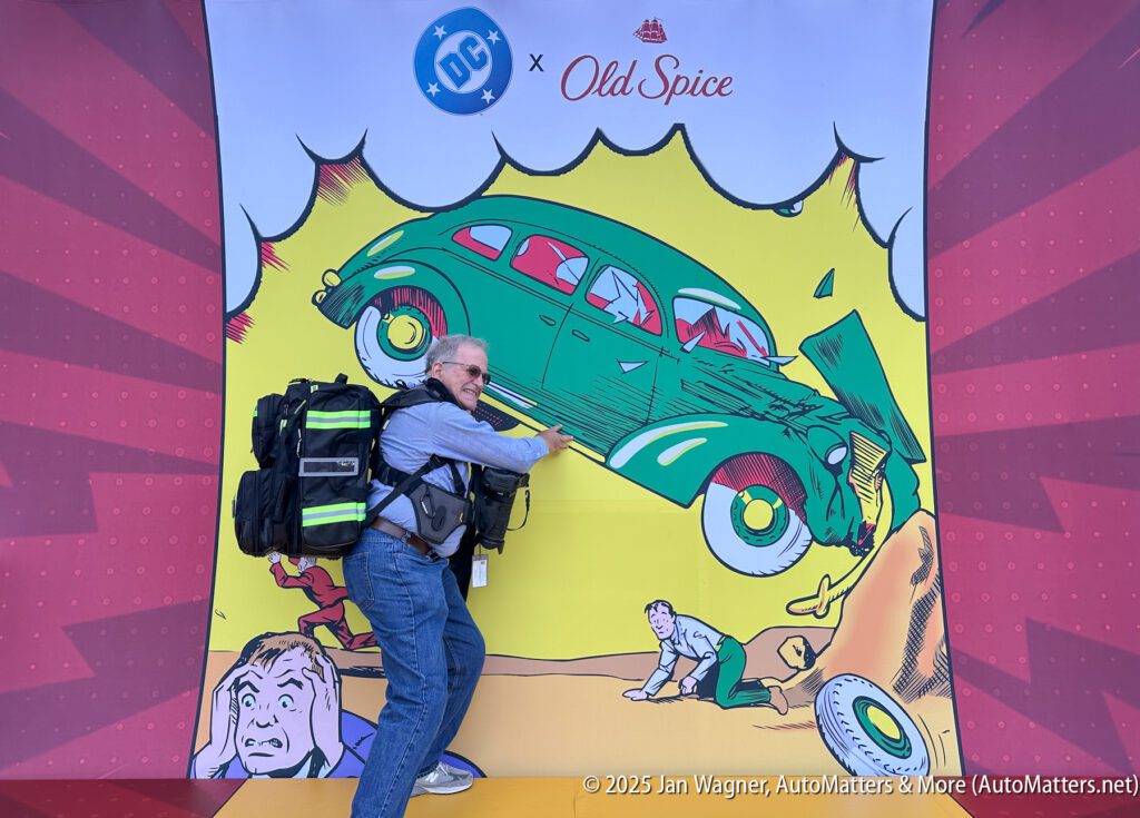 A man with a backpack poses in front of a comic-style backdrop showing a green car crashing through a wall, with DC and Old Spice logos above.