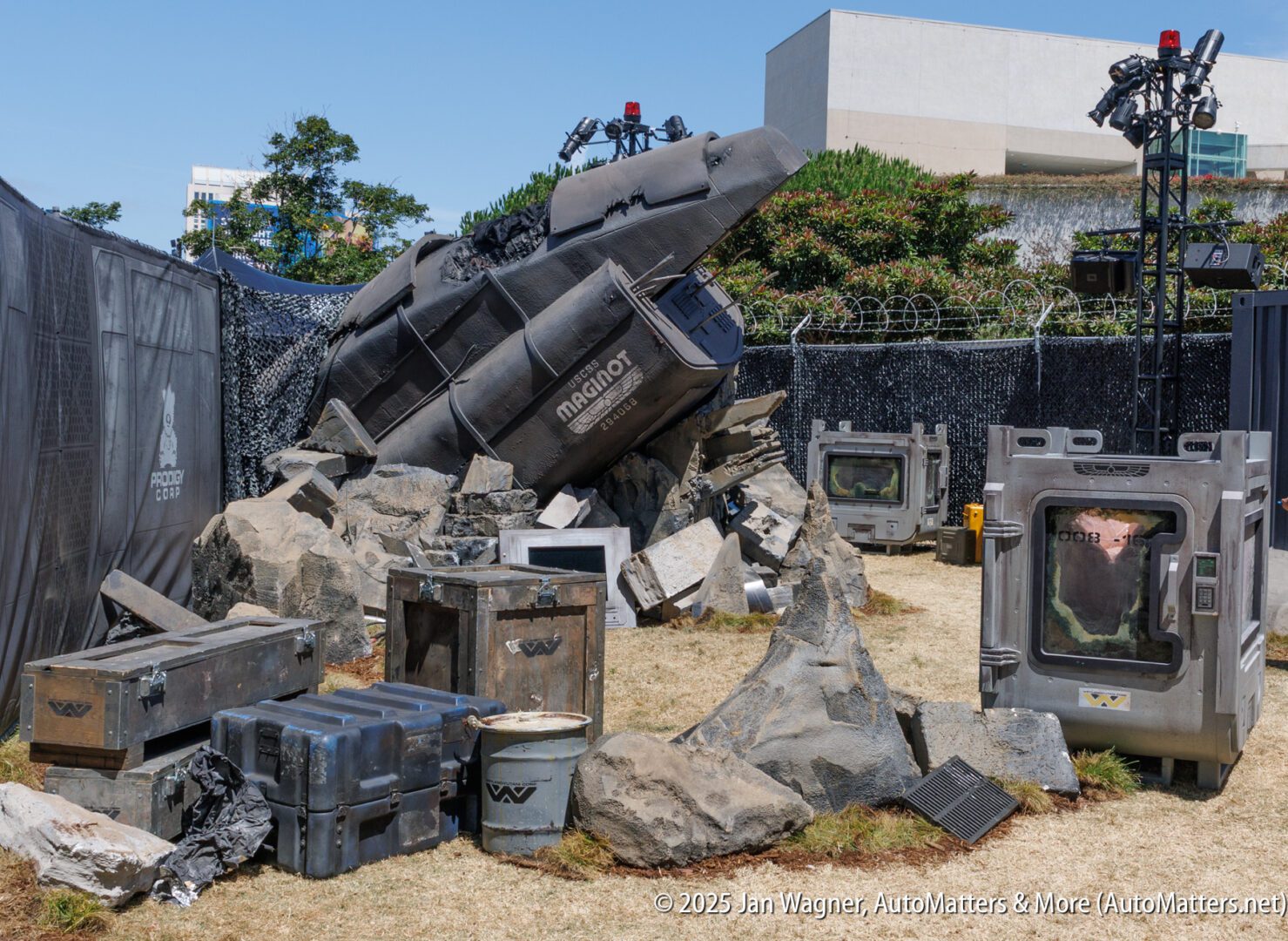 A crashed spaceship prop surrounded by rocks and sci-fi containers is set up outdoors, with buildings and equipment in the background.