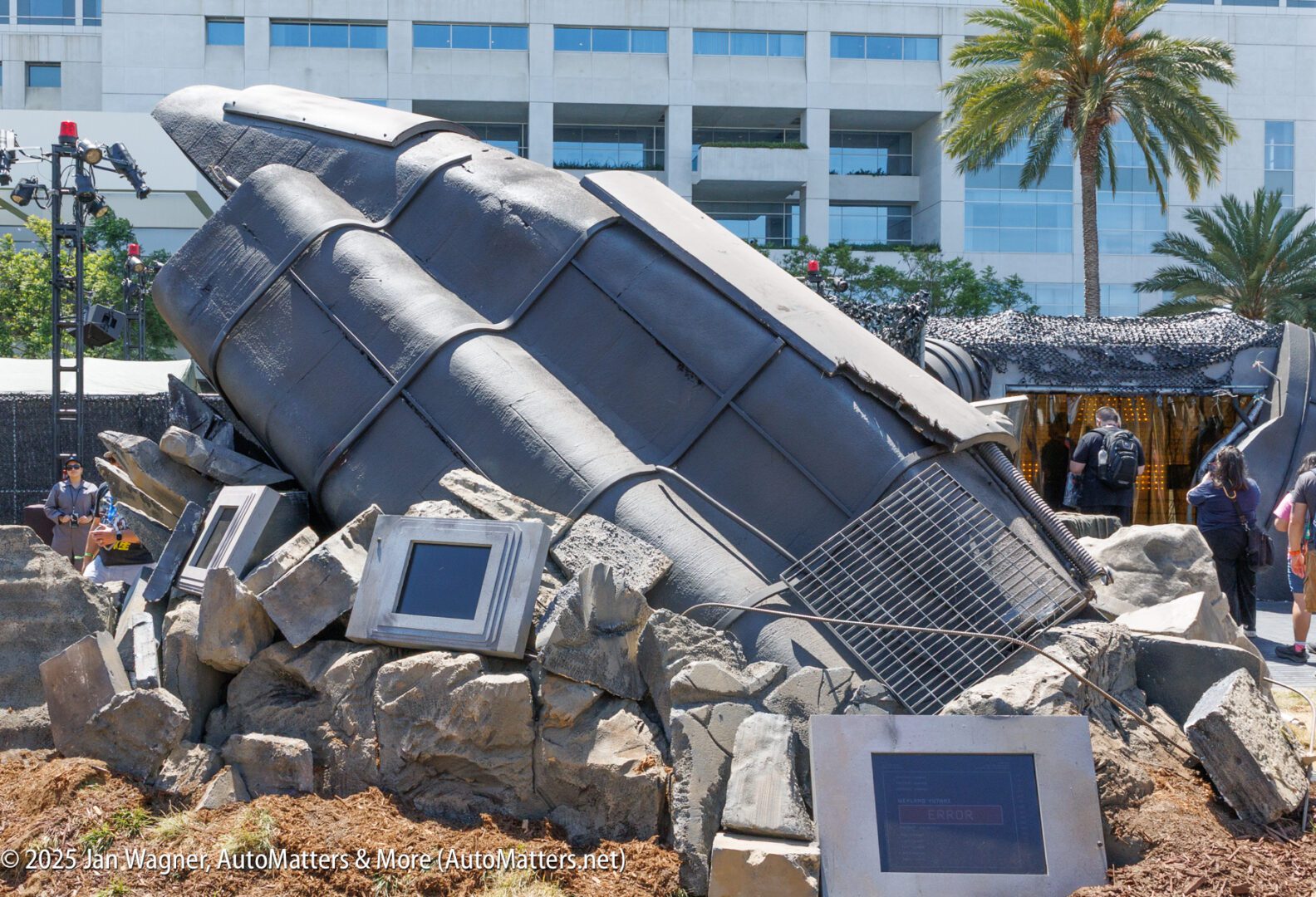 A large, metallic pod-like structure is partially embedded in rubble outdoors, surrounded by display screens and people, with a building and palm tree in the background.