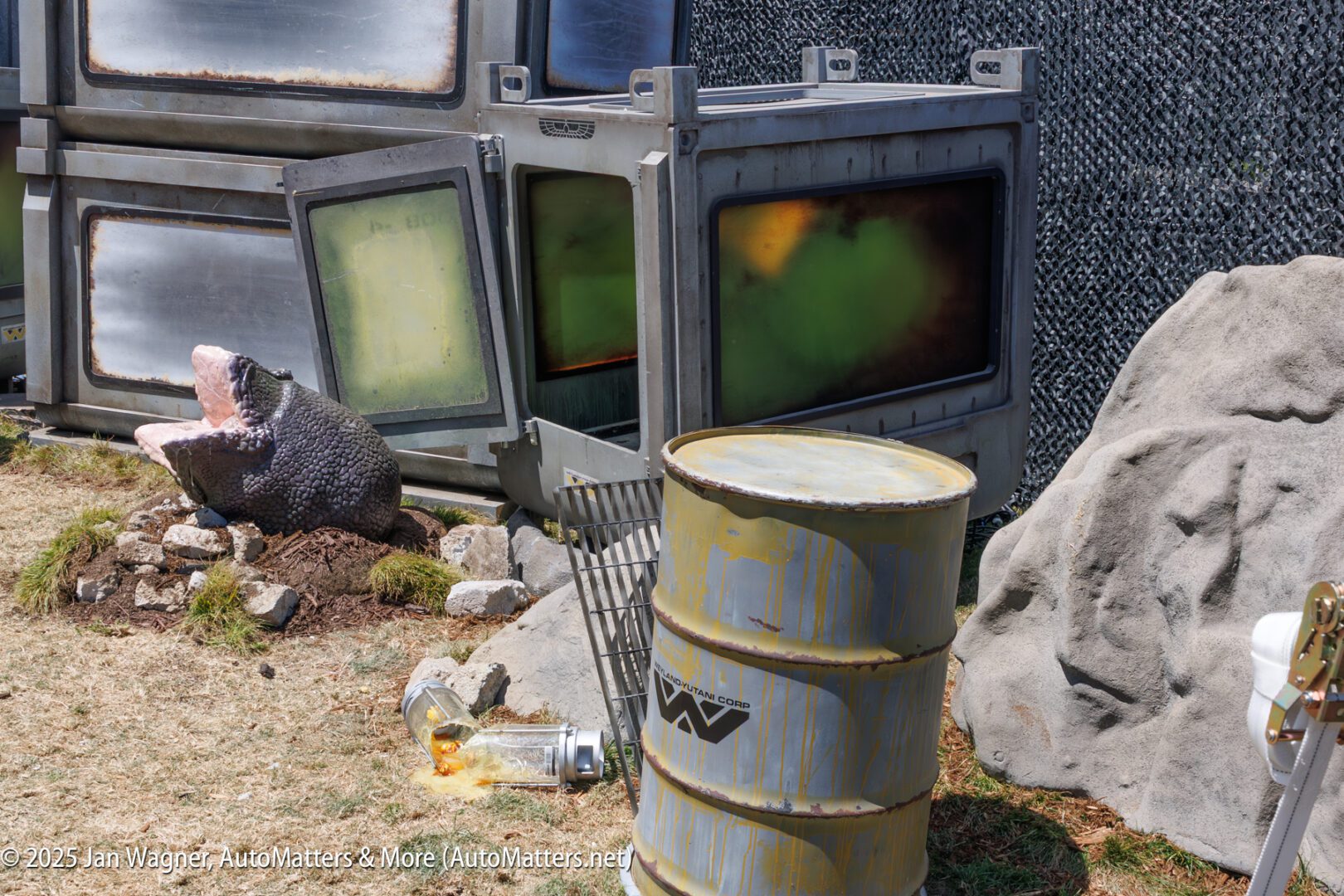 Two large industrial containers with green-tinted windows and a rusted barrel are surrounded by rocks, a broken shopping cart, and a large snake head prop on dry grass.