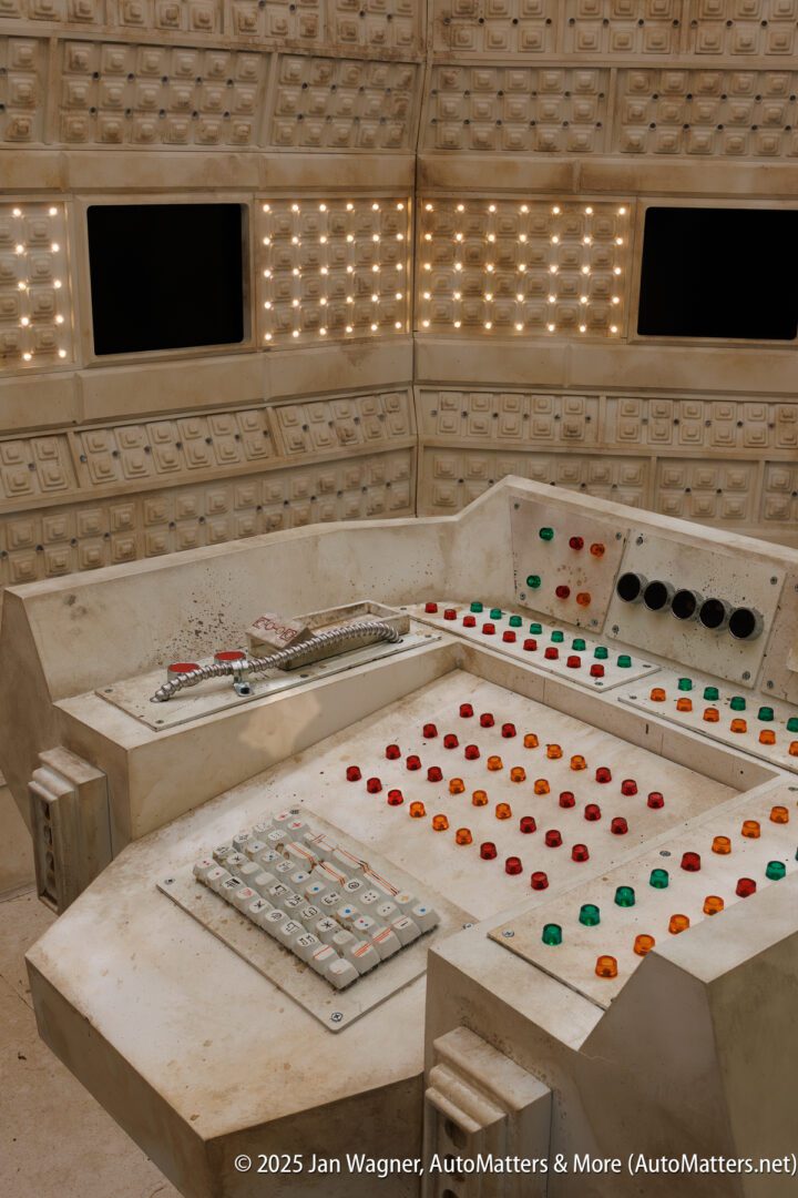 A retro-futuristic control panel with numerous buttons, switches, and indicator lights, set in front of large blank screens.