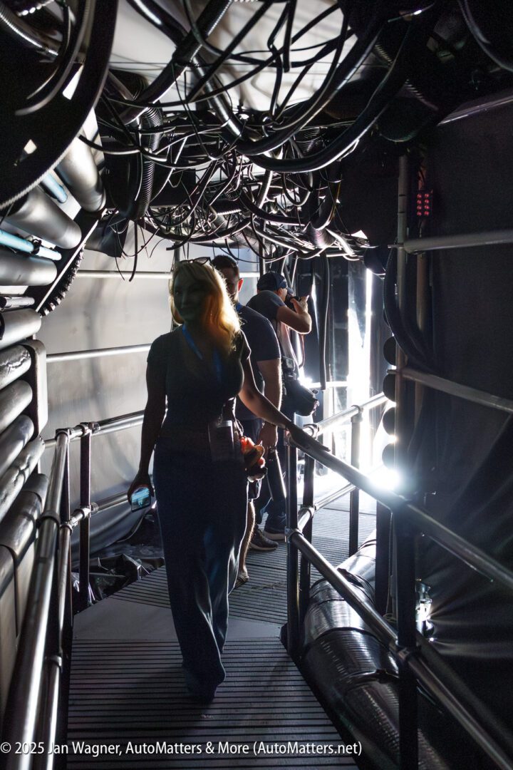 Two people walk through a dimly lit corridor with exposed cables and pipes overhead, illuminated by bright light at the far end.