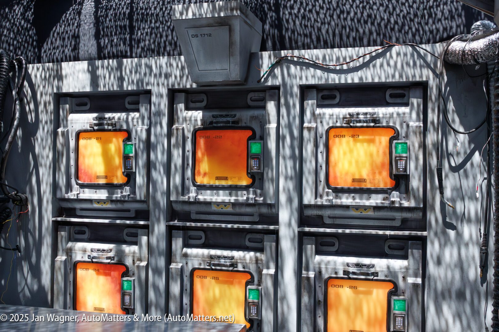 Six industrial-looking storage units with orange-lit windows and keypads are arranged in two rows of three, set against a background with cables and textured surfaces.